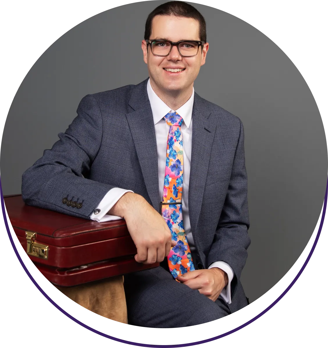 image design-3 Man in a suit with a colorful tie, sitting and resting his arm on a briefcase, smiles at the camera.