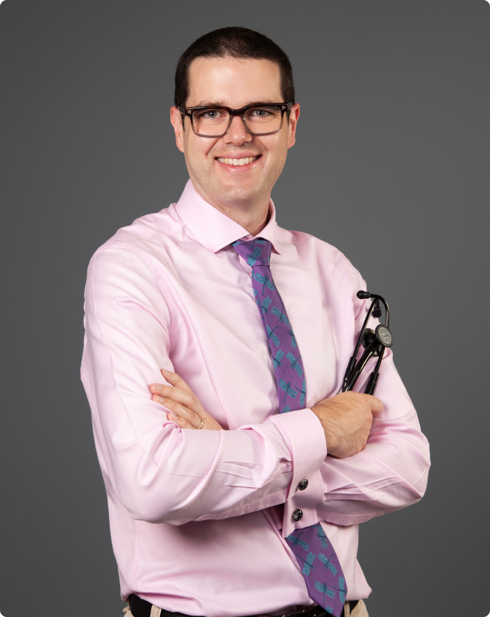rectangle_1108_1x A person wearing glasses, a pink shirt, and a patterned tie stands against a gray background, smiling with arms crossed and holding a stethoscope.